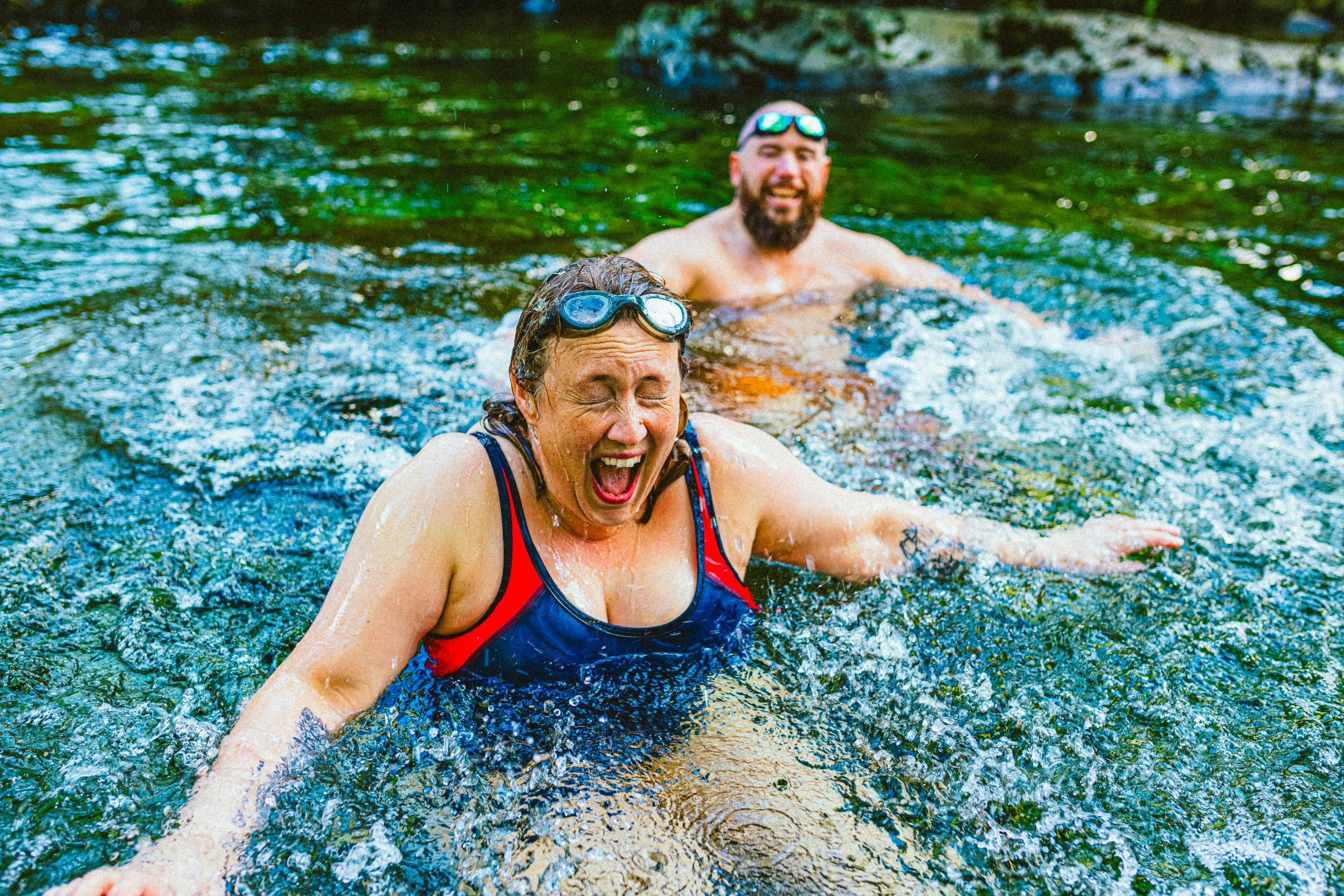 1492273951 Couple wild swimming in the Lake District, North West of England. They are in a river, enjoying time outdoors. The woman is laughing with her eyes closed and mouth open.
