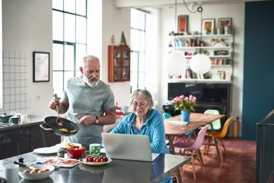 An older couple, one looking at a laptop and the other cooking.