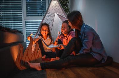 A mom with her two daughters camp inside their home due to the coronavirus restrictions and quarantine.They have pitched a tent along and have a fake campfire next to their tent. They are preparing marshmallows on campfire. They are making the best of ...