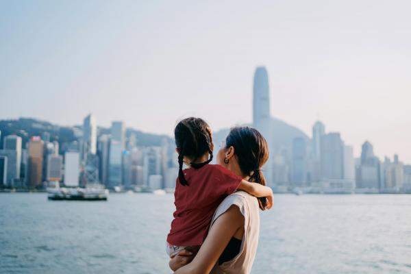 Loving young Asian mother embracing adorable little daughter in arms while looking over the city by the promenade of Victoria harbour. Enjoying the spectacular urban city skyline at sunset. Sharing intimate bonding time together