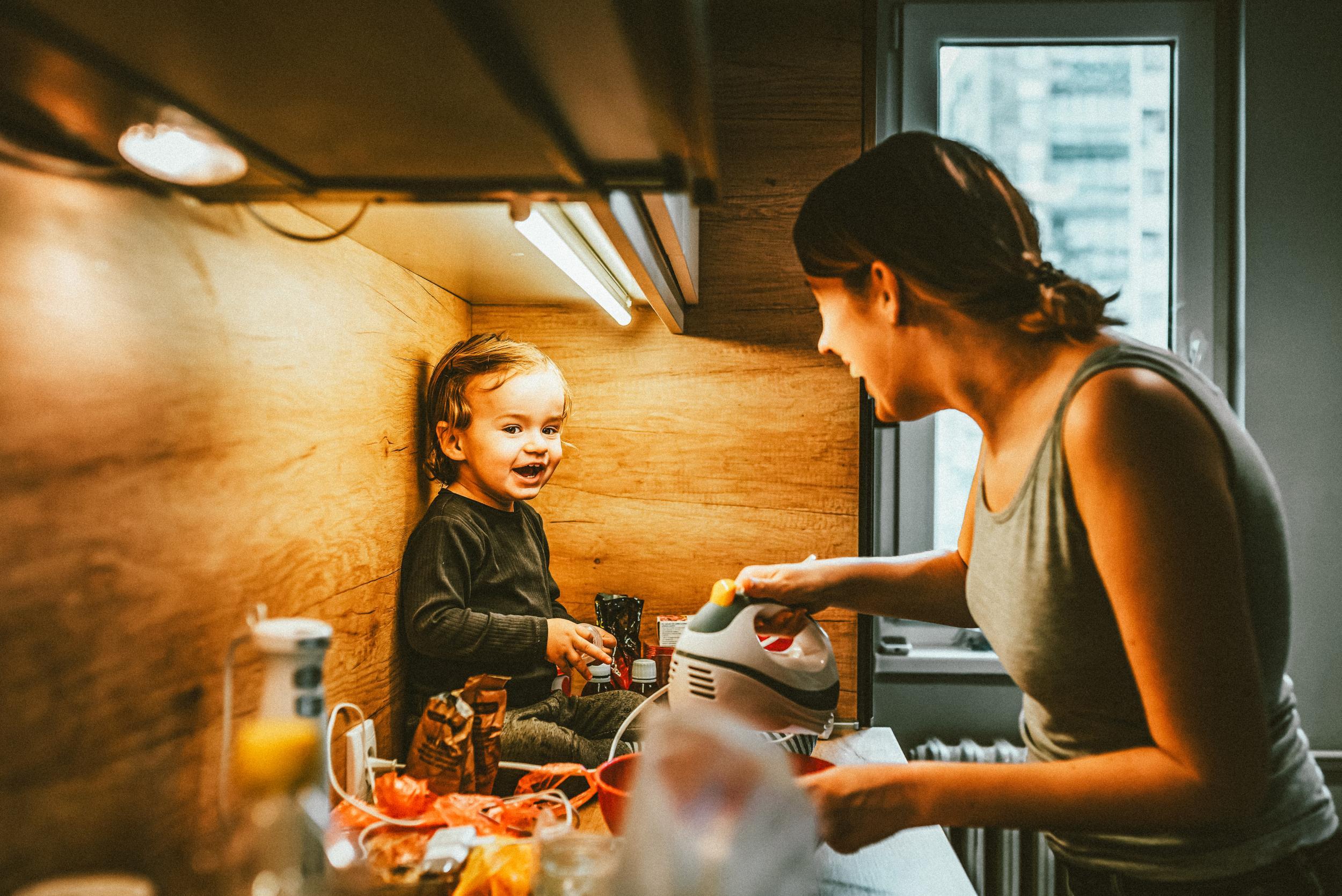 Family, food, healthy eating, cooking and people concept - happy mother working in kitchen with little baby boy at home kitchen. Mother with her baby son in the kitchen cooking together during the day. Smiling mother preparing dinner for her little chi...