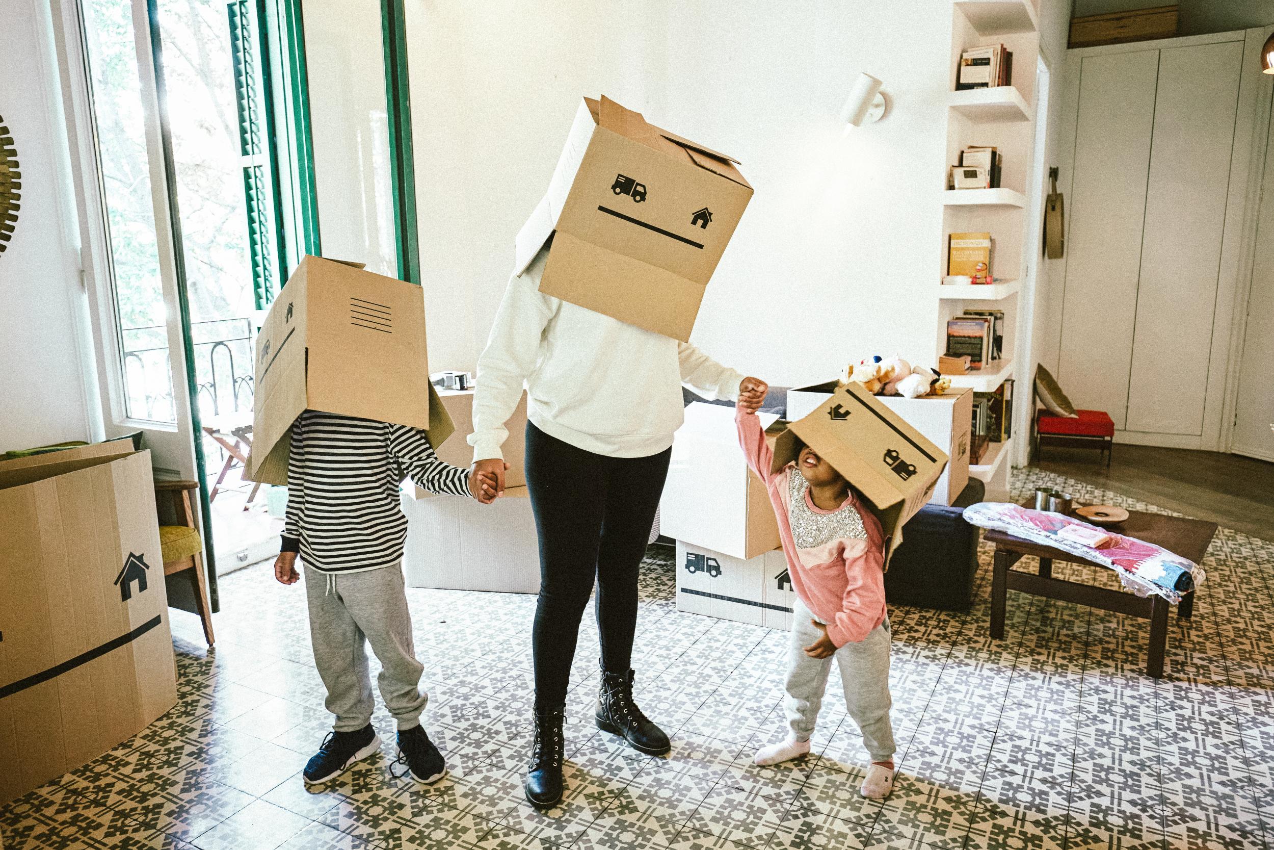 Full length view of Afro-Caribbean mother holding hands of young son and daughter as they take a break from moving to have fun with cardboard boxes.