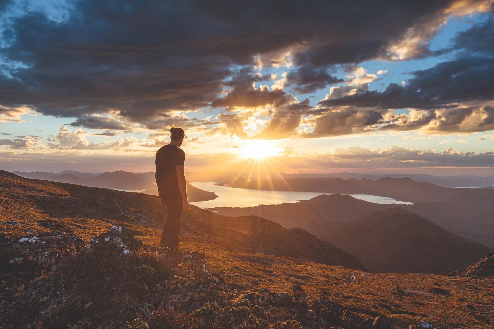 Man standing on cliff watching sunset over golden mountain range, Tasmania, Australia